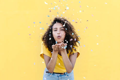 Young woman blowing multi colored confetti against yellow wall - RDGF00229