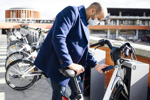 Man wearing protective face mask unlocking bicycle through smart phone on sunny day - PGF00199