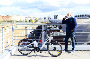 Man talking through smart phone while standing with hand in pockets on bridge during COVID-19 - PGF00212