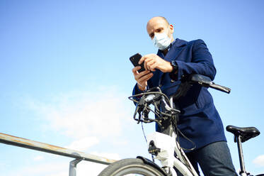 Mature man using smart phone while standing with bicycle against blue sky on sunny day during coronavirus - PGF00213
