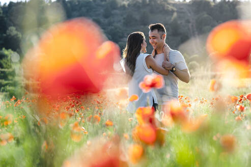 Glückliches romantisches Paar lächelt sich gegenseitig an, während es auf einem Mohnblumenfeld steht - JMPF00587