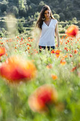 Junge Frau steht inmitten von Mohnpflanzen auf einem Feld im Frühling - JMPF00590
