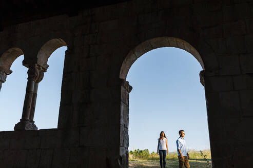 Young couple standing on field seen through arch at Roman church - JMPF00593