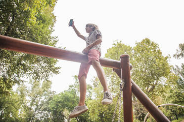 Junge macht Selfie, während er auf einem Spielgerät im Park sitzt - AJOF00577