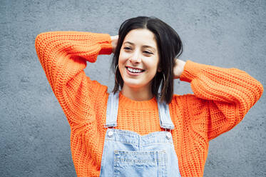 Lächelnde Frau in orangefarbenem Pullover schaut weg, während sie an einer grauen Wand steht - PGF00216