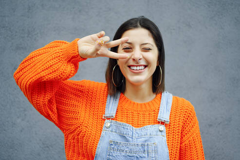 Young woman showing peace sign while standing against gray wall - PGF00218