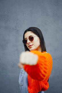 Selbstbewusste Frau mit Sonnenbrille zeigt Schlag, während sie gegen eine graue Wand steht - PGF00219