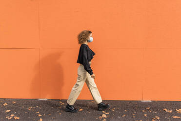 Junge Frau mit Gesichtsmaske läuft gegen eine orangefarbene Wand - MEF00001