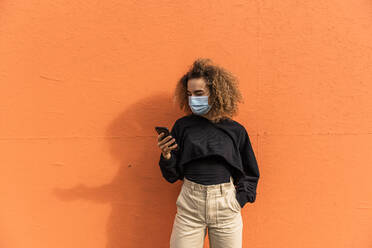 Young woman wearing face mask using mobile phone while standing against orange wall - MEF00005