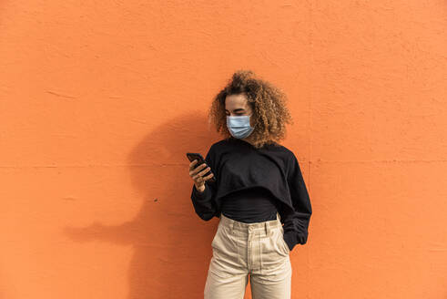 Junge Frau mit Gesichtsmaske, die ein Mobiltelefon benutzt, während sie an einer orangefarbenen Wand steht - MEF00005