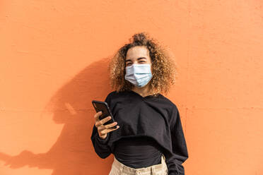 Junge Frau mit Gesichtsmaske, die wegschaut, während sie ein Mobiltelefon an einer orangefarbenen Wand benutzt - MEF00006