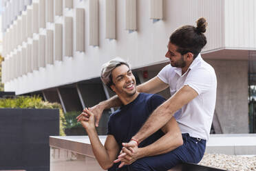 Lächelnder Mann, der seinen schwulen Partner ansieht, während er an einem Gebäude in der Stadt sitzt - PNAF00135