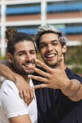 Smiling man showing engagement ring while standing by male partner in park - PNAF00184