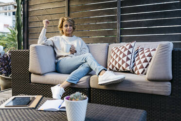 Blonde Frau schaut weg und hält eine Kaffeetasse auf dem Sofa im Dachgarten - JRFF04912
