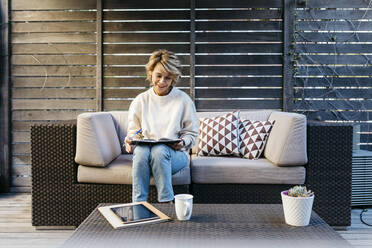 Smiling woman writing in diary while sitting on sofa at rooftop garden - JRFF04913