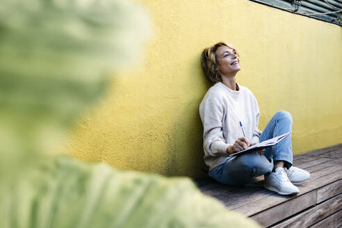 Thoughtful smiling woman sitting with diary against yellow wall on building terrace - JRFF04919