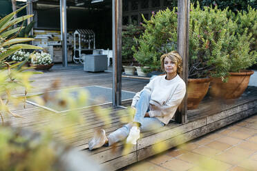Blond woman contemplating while sitting on wooden floor at rooftop garden - JRFF04924