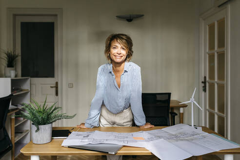 Smiling female architect leaning on desk while standing in office - JRFF04955