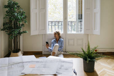 Geschäftsfrau, die ein Smartphone benutzt, während sie auf dem Boden vor dem Fenster im Büro sitzt - JRFF04957