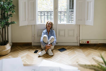 Thoughtful businesswoman sitting on floor against window in office - JRFF04960