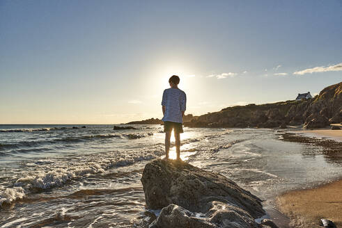 Boy admiring sea view while standing on rock at beach - JEDF00338