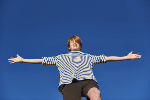 Boy standing with arms outstretched against clear sky - JEDF00340