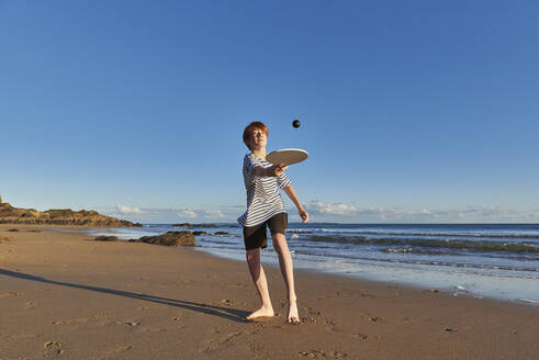 Boy playing with ball and racket while standing at beach - JEDF00341