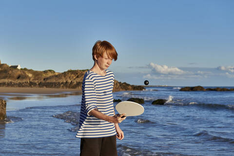 Boy playing with ball and racket while standing at against sea stock photo