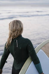 Ältere Frau hält Paddleboard und schaut auf das Meer, während sie in der Morgendämmerung steht - EGAF01036