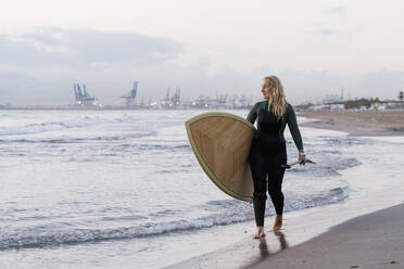 Blond woman holding paddleboard walking at beach during dawn - EGAF01037