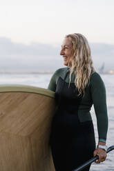 Blonde Frau lacht, während sie ein Paddleboard am Strand in der Morgendämmerung hält - EGAF01038