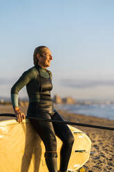 Frau lacht und schaut weg, während sie sich am Strand in der Morgendämmerung auf ein Paddleboard stützt - EGAF01054