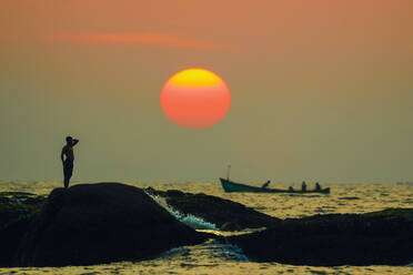 Mann auf Gezeitenfelsen und Fischerboot bei Sonnenuntergang am wunderschönen, unberührten Kizhunna Beach, südlich von Kannur, Kannur, Kerala, Indien, Asien - RHPLF17797