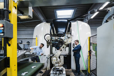 Businessman explaining male colleague about machinery while standing in factory - DIGF13119