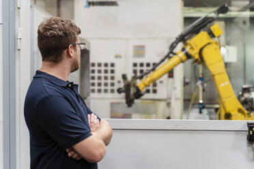 Männlicher Fabrikarbeiter mit verschränkten Armen, der einen Roboterarm durch ein Fenster betrachtet, während er in einer Fabrik steht - DIGF13144