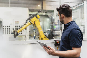 Young male worker looking at robotic arm while holding digital tablet in factory - DIGF13147