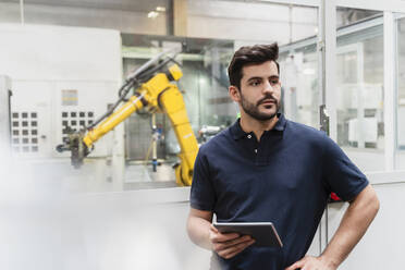 Männlicher Arbeiter, der wegschaut und ein digitales Tablet hält, steht in einer Fabrik - DIGF13150