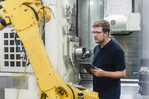 Männlicher Arbeiter, der einen Roboterarm untersucht, während er ein digitales Tablet in einer Fabrik hält - DIGF13152