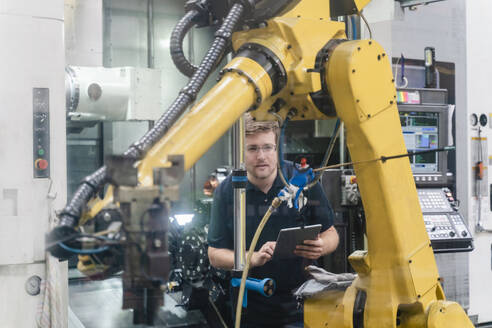 Männlicher Arbeiter, der einen Roboterarm analysiert, während er ein digitales Tablet in einer Fabrik hält - DIGF13153