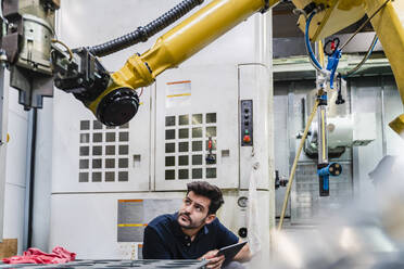 Junge männliche Arbeitskraft mit digitalem Tablet, die einen Roboterarm in einer Fabrik untersucht - DIGF13157