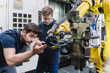 Male coworkers examining robotic arm while standing in manufacturing factory - DIGF13160