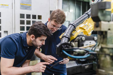 Männliche Mitarbeiter, die einen Roboterarm untersuchen, während sie ein digitales Tablet in einer Produktionsstätte benutzen - DIGF13161