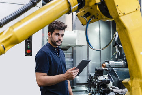 Männlicher Arbeiter, der ein digitales Tablet hält und wegschaut, während er in einer Fabrik steht - DIGF13166