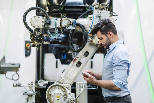 Männlicher Unternehmer, der ein Maschinenteil untersucht, während er an einem Roboterarm in der Industrie steht - DIGF13193