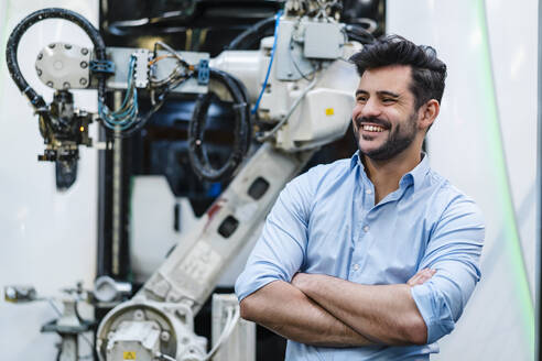 Smiling male entrepreneur with arms crossed looking away while standing by robotic arm at industry - DIGF13199