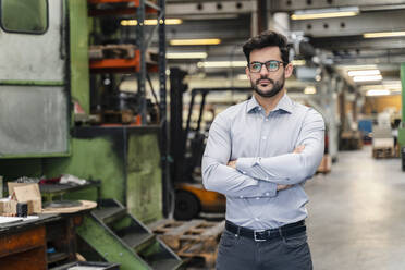 Confident businessman with arms crossed standing at manufacturing industry - DIGF13226