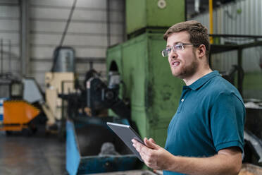 Male worker looking away while holding digital tablet standing in factory - DIGF13228