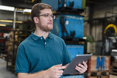 Nachdenklicher männlicher Arbeiter, der ein digitales Tablet hält, während er in einer Fabrik steht - DIGF13230