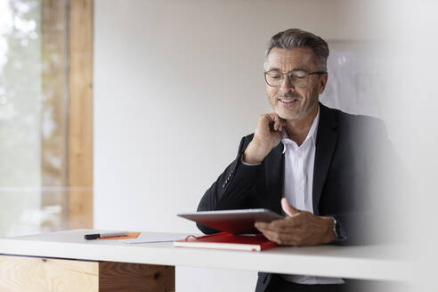 Lächelnder Geschäftsmann mit Hand am Kinn, der zu Hause ein digitales Tablet benutzt - PESF02176