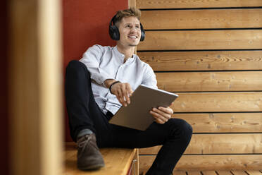 Man wearing headphones with digital tablet looking away while sitting at home - PESF02210